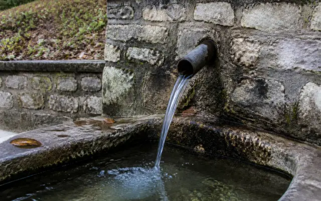 O fântână amenajată din piatră de unde curge apă printr-o țeavă metalică într-un bazin de piatră plin cu apă curată