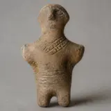 O figurină antropomorfă din lut ars, cu aspect arhaic, prezentând detalii incizate la nivelul gâtului (asemănătoare unor coliere) și taliei (ca o centură)