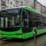Un autobuz electric modern de culoare verde aprins oprit într-o stație pe marginea unei străzi urbane umede, cu clădiri de apartamente de tip bloc în fundal