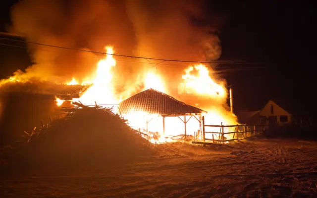 A large house engulfed in intense orange flames and thick smoke at night during a structural fire